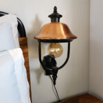 Pair of restored vintage wall sconces with aged copper domed shades, rustic patina finish, black metal necks and glowing Edison bulbs mounted on a wall.